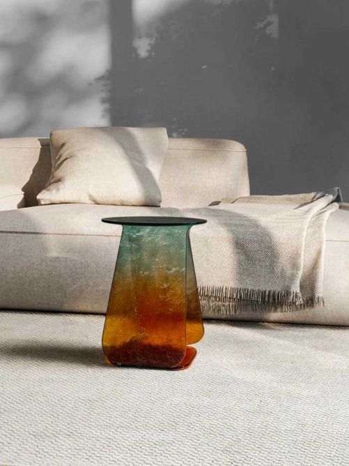 Skulpturaler Tisch aus verschmolzenem Murano-Glas von Mademoiselle Jo. Die belgische Designerin Joan Bebronne überträgt ihre Erfahrung aus Architektur sowie Mode- und Textildesign in zeitgenössische Möbel und Wohnobjekte. Ihre reduzierte Formensprache verbindet sie mit hochwertigen Materialien und präziser Verarbeitung. Die limitierten Editionen entstehen in aufwendiger Handarbeit in den Werkstätten von Murano: Mehrere miteinander verbundene Glasschichten erzeugen Oberflächen, auf denen Licht, Farbe und Tiefe stets neu zusammenspielen. www.mademoisellejo.com