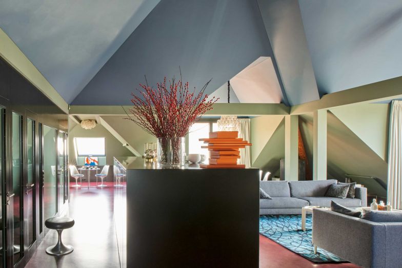 Die Nordseite des Lofts: Eine Wand aus Glas und MDF mit Eingangstür, Dusche und Toilette. Als Bodenbelag: rotes Linoleum, Wände, Decken und Balken in Grün und Blau. Ein gross dimensioniertes Sideboard dient als Stauraum für Kleider und Geschirr und als Raumtrenner zwischen dem Eingangs- und dem Wohnbereich.