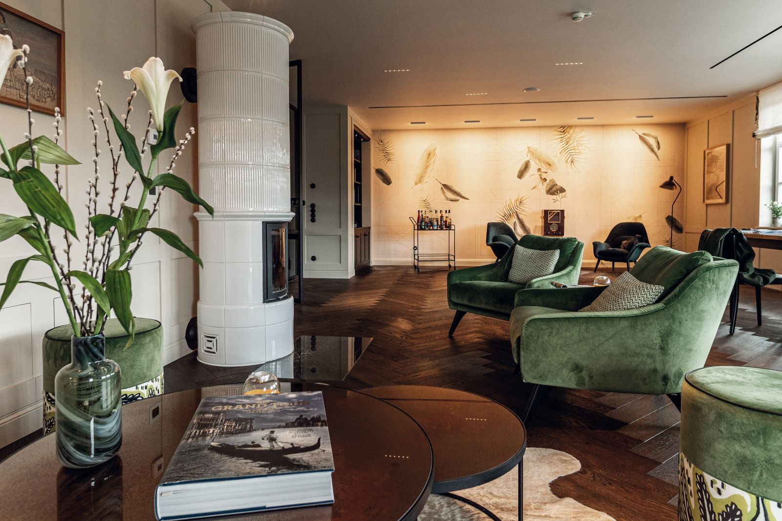 Im Erdgeschoss liegt der grosse Wohn-/Essbereich. Hier befindet sich auch eine kleine Bibliothek mit Barwagen. Farblich abgestimmte Polstermöbel laden zum Lesen und Verweilen ein. Hingucker ist die Tapete «Phytogenesis» mit Blättermotiven von Wall & Deco. Die davor stehenden Loungesessel «Swoon» sind von Fredericia. Die grünen Sessel im Bild vorne sind von IP Design, Modell «Flow Lounge Chair».