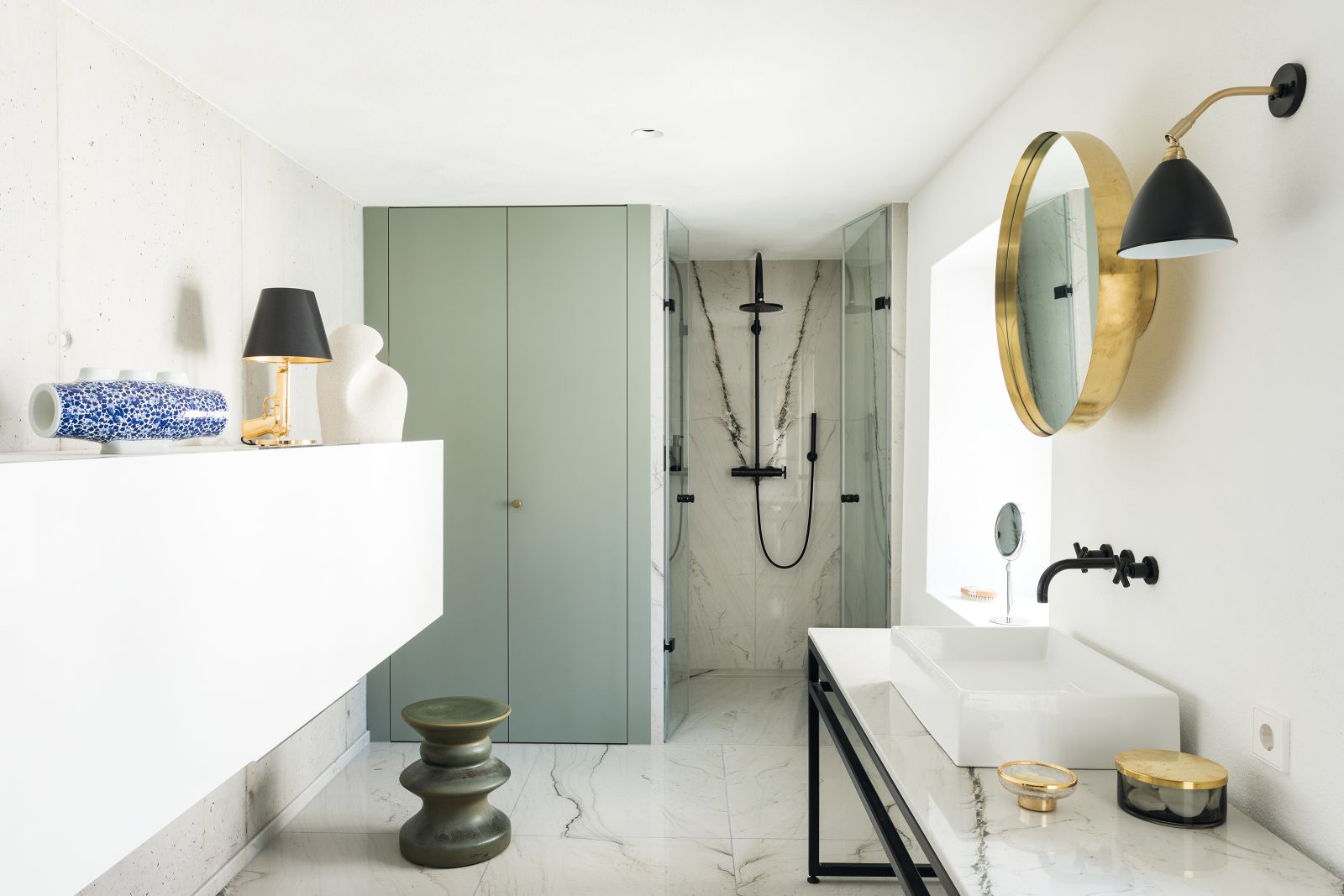 Im Elternbad kombiniert die Innenarchitektin edle Accessoires aus Messing mit hellem Naturstein und schwarzen Armaturen. Spiegel «Darkly» von Nick Ross Studio für Menu. Armaturen, Dornbracht. Hocker, Zanotta. Leuchte «Bestlite BL7» von Robert Dudley Best für Gubi. Vase «Delft Blue No. 4» aus Porzellan von Marcel Wanders für Moooi.