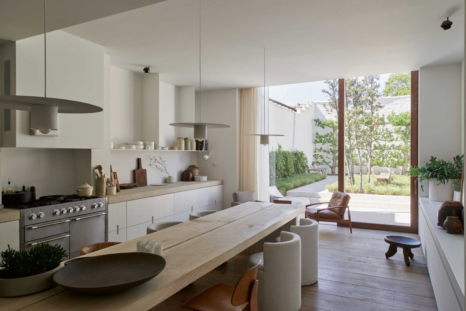 Die Wohnküche liess Paul Vanrunxt in mattem Weiss streichen und kombiniert dieses mit beigem Kalkstein. Das helle Ambiente verleiht dem vielschichtigen Interieur einen uniformen Look. Der Esstisch aus massivem Pappelholz ist ein Eigenentwurf, ebenso die Porzellanleuchten darüber.