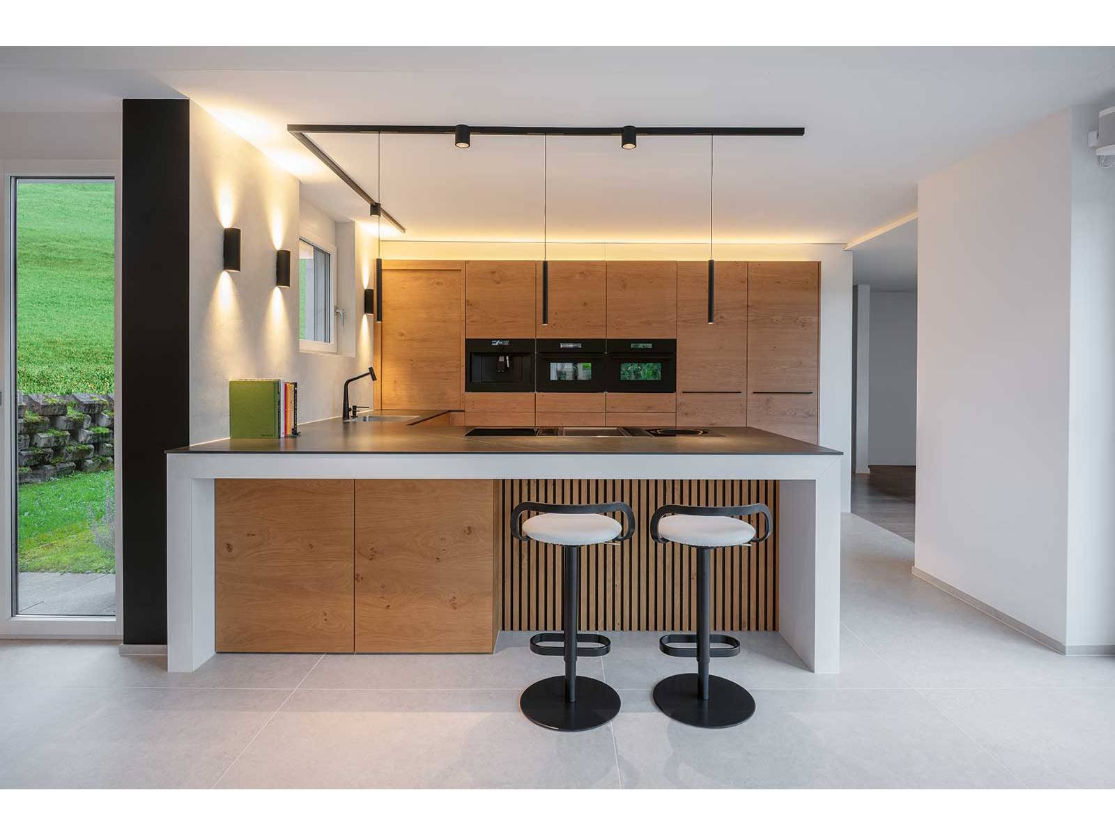 In warmer Eiche und mit klaren Linien präsentiert sich diese offene Küche. Die weisse Insel mit feiner Holzverkleidung und zwei bequemen Barhockern bildet ein einladendes Zentrum. Präzise schwarze Akzente – Abdeckung, Griffe, Armaturen, Spotlights – verleihen Struktur, während das Lichtkonzept Tiefe und Funktionalität schafft. ELBAU.