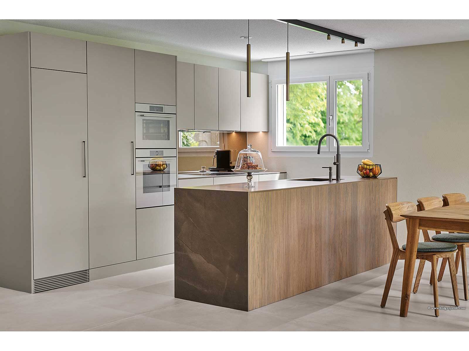 Edle Materialien und harmonische Farbtöne prägen ­diesen Küchenumbau. Eine grosszügige Insel mit reliefierter Eichenverkleidung verleiht Wärme und natürliche Struktur, die Keramik-Arbeitsplatte mit feiner Marmorierung fügt Eleganz und Robustheit hinzu. Der bronzefarbene Parsol­spiegel an der Rückwand reflektiert Licht und schafft Tiefe, schwarze Armaturen ergänzen das durchdachte Gesamt­konzept. BRUNNER KÜCHEN.