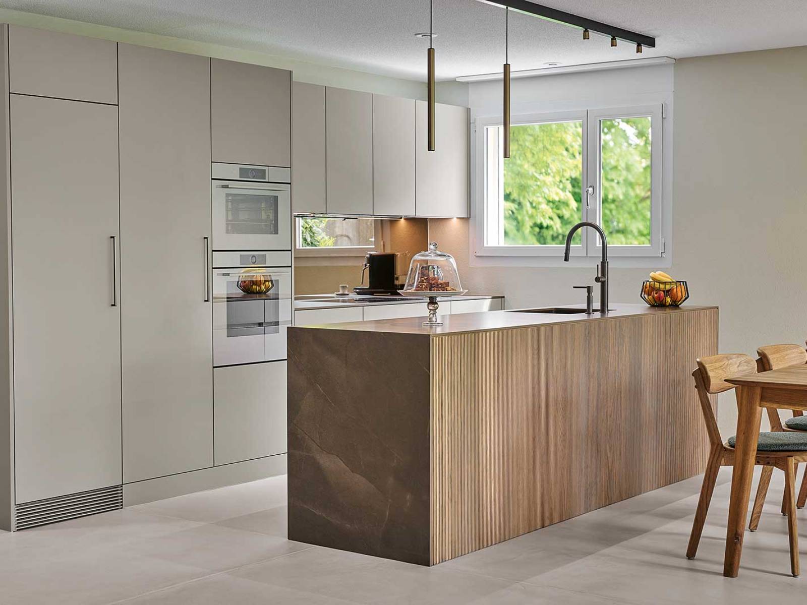 Edle Materialien und harmonische Farbtöne prägen ­diesen Küchenumbau. Eine grosszügige Insel mit reliefierter Eichenverkleidung verleiht Wärme und natürliche Struktur, die Keramik-Arbeitsplatte mit feiner Marmorierung fügt Eleganz und Robustheit hinzu. Der bronzefarbene Parsol­spiegel an der Rückwand reflektiert Licht und schafft Tiefe, schwarze Armaturen ergänzen das durchdachte Gesamt­konzept. BRUNNER KÜCHEN.