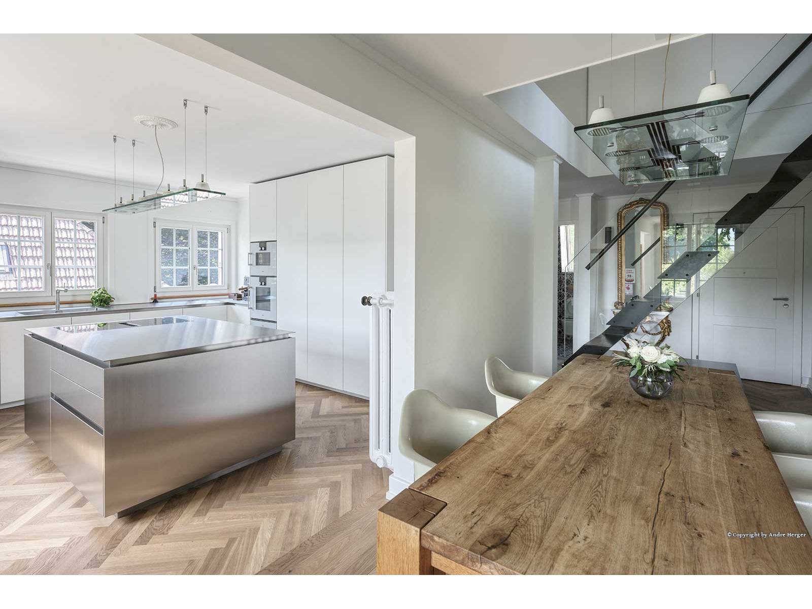 Diese Küche lebt von puristischer Präzision. Raum­hohe Schränke bilden den ruhigen Hintergrund zur ­markanten Edelstahlinsel, deren grosszügiges Kochfeld mit Muldenlüftung genug Platz für das Mise en place lässt. Edelstahl erstreckt sich flächendeckend über die ­Küchenzeile und integriert nahtlos Spüle mit Kräuter­garten. Hochwertige Geräte gewährleisten makellose Funktionalität in heller, offener Raumgestaltung. Foto André Herger. OREA.