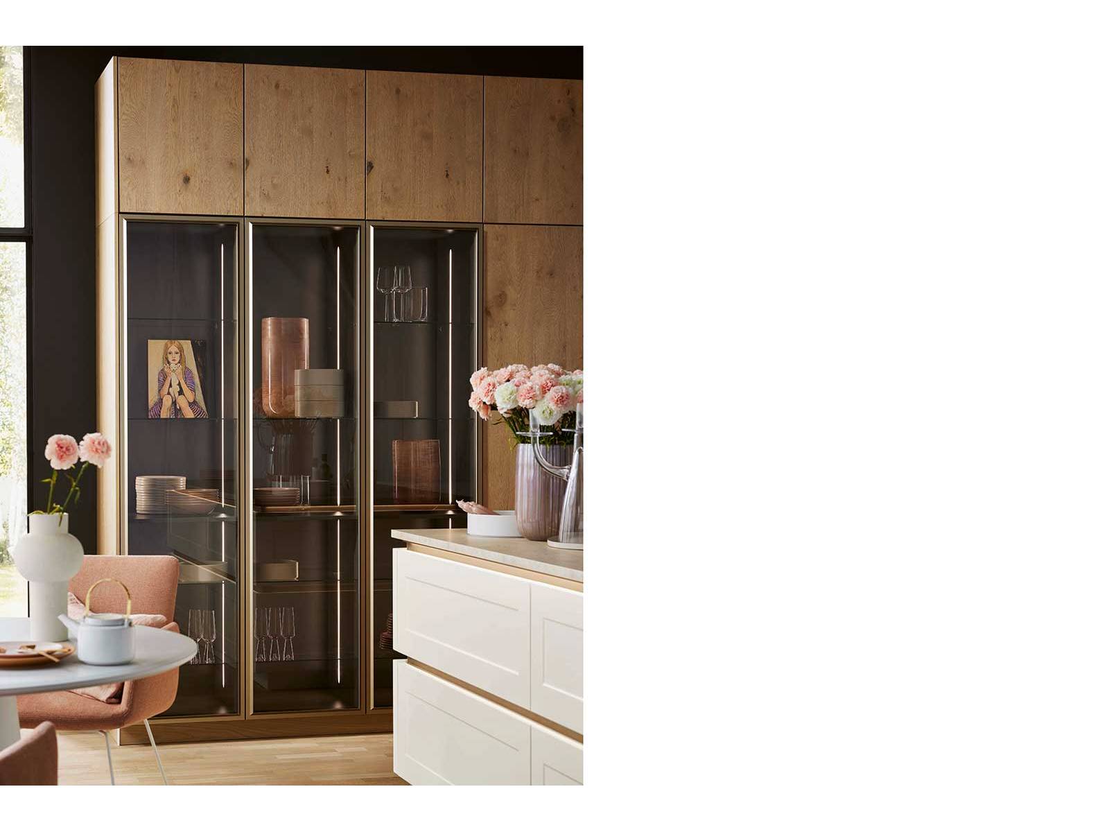 Ein eleganter Landhauslook entsteht bei dieser Küche durch die Kombination der grifflosen Land­hausfronten mit den Schränken «Tavola» in Eiche Pinot sowie den Griffspuren in Manganbronze, die sich ­harmonisch im Rahmen der Vitrine widerspiegeln. NOLTE KÜCHEN.