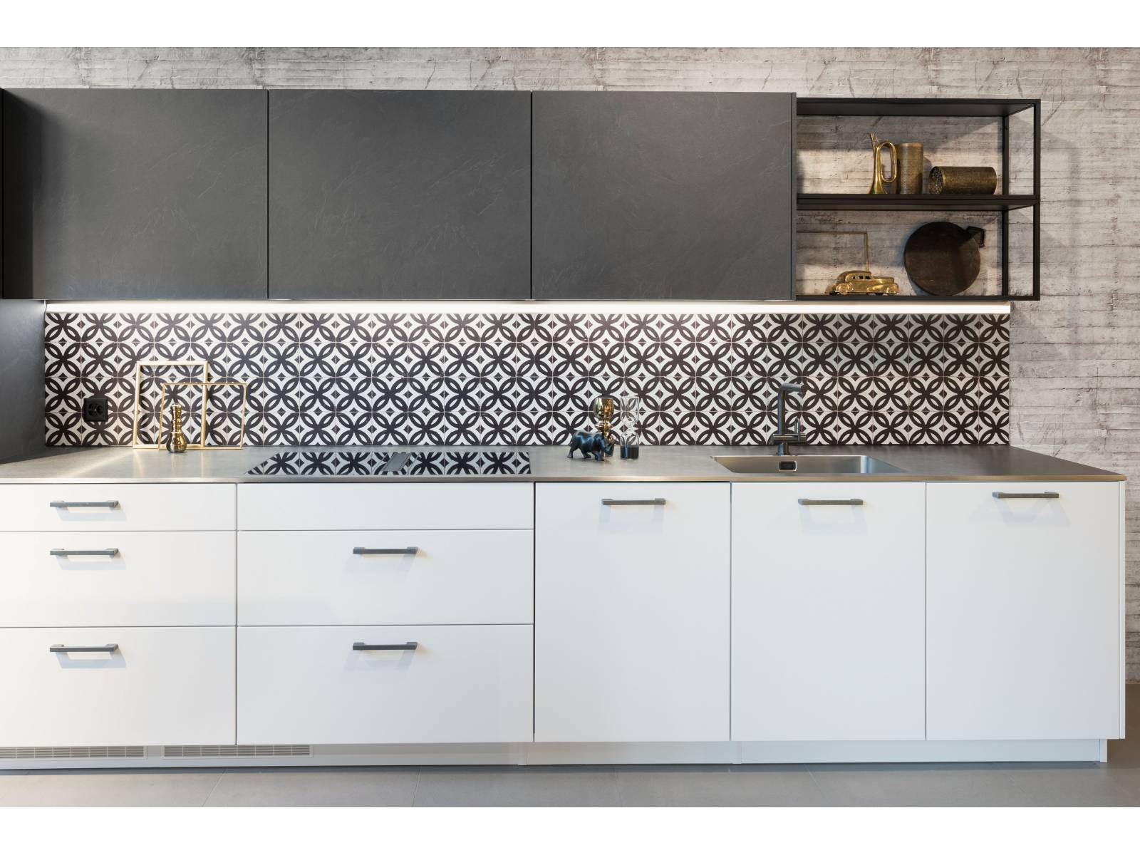 Sie liegen wieder im Trend – Ornamente, die bei dieser Küche die Rückwand zieren und so die Strenge der weissen und anthrazitfarbigen Küchenelemente auflockern. Ein Lichtband unter den Hochschränken sorgt dabei nicht nur für genügend Helligkeit, sondern setzt das Muster gekonnt in Szene. Sanitas Troesch.