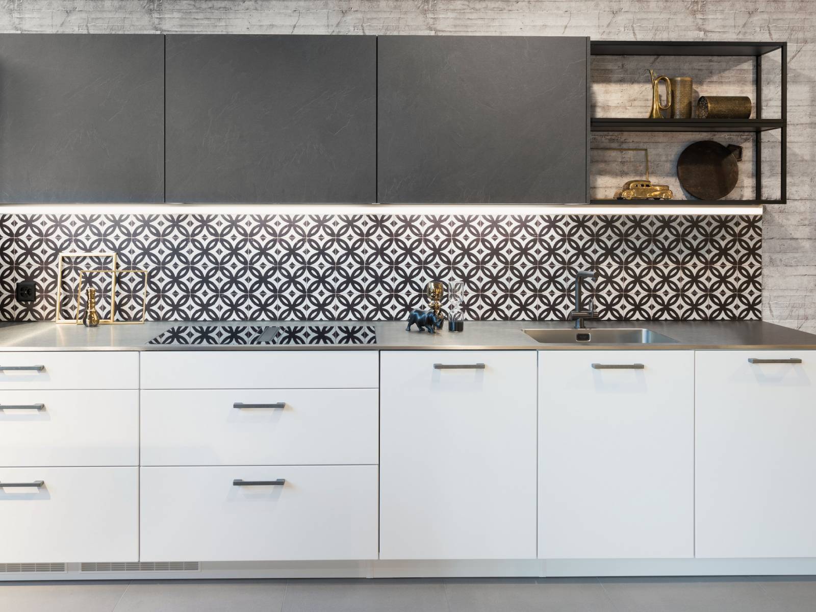 Sie liegen wieder im Trend – Ornamente, die bei dieser Küche die Rückwand zieren und so die Strenge der weissen und anthrazitfarbigen Küchenelemente auflockern. Ein Lichtband unter den Hochschränken sorgt dabei nicht nur für genügend Helligkeit, sondern setzt das Muster gekonnt in Szene. Sanitas Troesch.