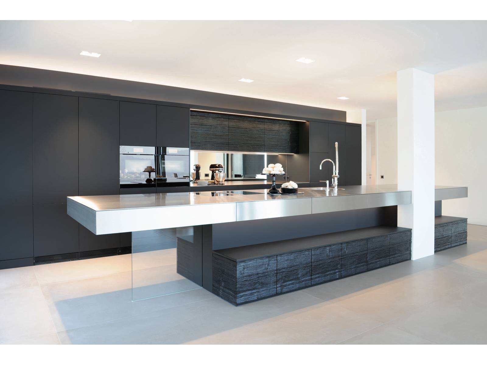 Trotz der dunklen Fronten und dem Einsatz von Strukturholz scheint die langgezogene Kücheninsel mit Chromstahlfronten regelrecht zu schweben. Eine statische Glaswand gibt sowohl dem Küchenblock als auch dem Tisch Halt, der zwischen den Hochschränken integrierte Spiegel verleiht Raumtiefe. Brunner.