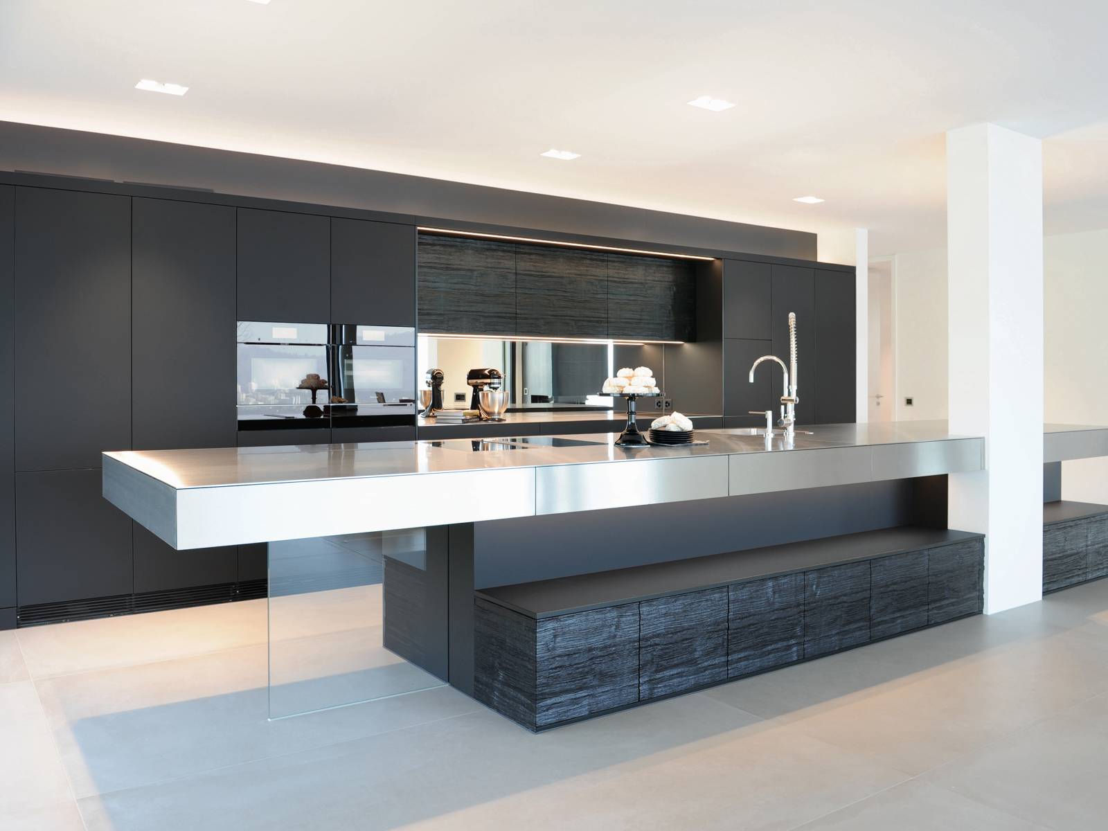 Trotz der dunklen Fronten und dem Einsatz von Strukturholz scheint die langgezogene Kücheninsel mit Chromstahlfronten regelrecht zu schweben. Eine statische Glaswand gibt sowohl dem Küchenblock als auch dem Tisch Halt, der zwischen den Hochschränken integrierte Spiegel verleiht Raumtiefe. Brunner.