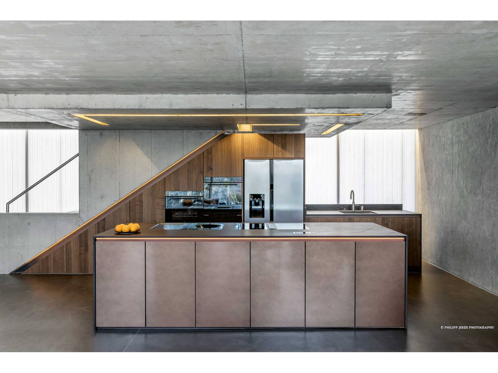 Es ist das Zusammenspiel der Materialien, das diese Küche zu einem Blickfang macht: Die gerillten Fronten aus Echtholzfurnier har­­­monieren mit denen der Kücheninsel in speziellem Metalldesign. Die dunkelbraune Arbeitsplatte ist mit goldenen Adern durchzogen, was bei der Beleuchtung farblich aufgenommen wurde. Gleich­­zeitig bildet die Küche einen spannenden Kontrast zum Sichtbeton und strahlt Wärme und Gemütlichkeit aus. SANITAS TROESCH.