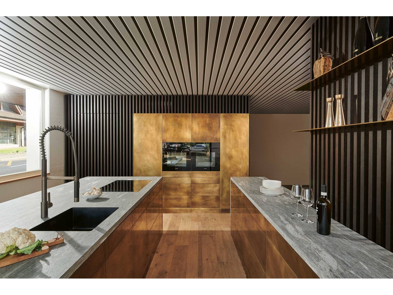 Ein wahres Goldstück ist die Küche mit Fronten aus handgewirbeltem Messing. In der Kombination mit Valser Quarzit entsteht eine spannende Verbindung, die durch das puristische Design nur noch verstärkt wird. Raffinierte Details, wie die integrierte Reduit-Türe aus Räuchereiche, ergänzen das Konzept. Foto: www.holgerjacob.com. BRUNNER KÜCHEN.