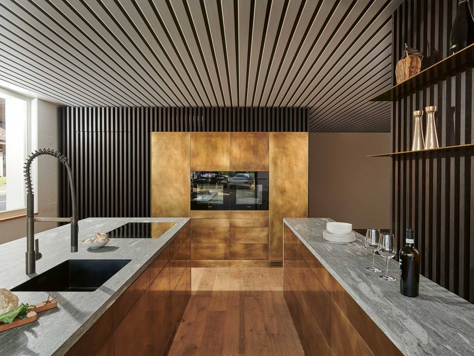 Ein wahres Goldstück ist die Küche mit Fronten aus handgewirbeltem Messing. In der Kombination mit Valser Quarzit entsteht eine spannende Verbindung, die durch das puristische Design nur noch verstärkt wird. Raffinierte Details, wie die integrierte Reduit-Türe aus Räuchereiche, ergänzen das Konzept. Foto: www.holgerjacob.com. BRUNNER KÜCHEN.