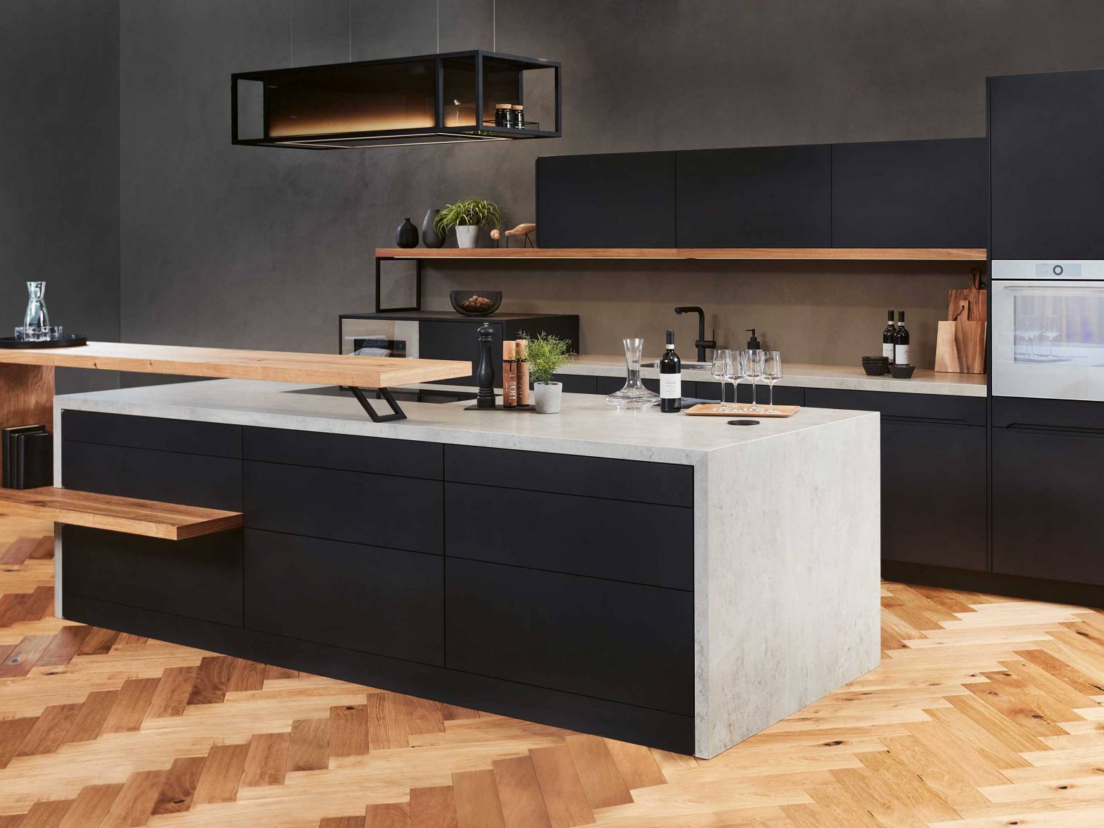 Die Küche der hauseigenen Linie «Infinity» spielt mit offenen und geschlossenen Elementen. Die schwarzen Hängeschränke werden von einem Regalboden aus Holz getragen, der in Form einer Spange vor der Kücheninsel seine Wiederholung findet. Eine betongraue Arbeitsplatte vollendet das neue Modell, das ganz auf Wunsch und nach Mass gefertigt wird. Foto: Studiojeker GmbH. SABAG.