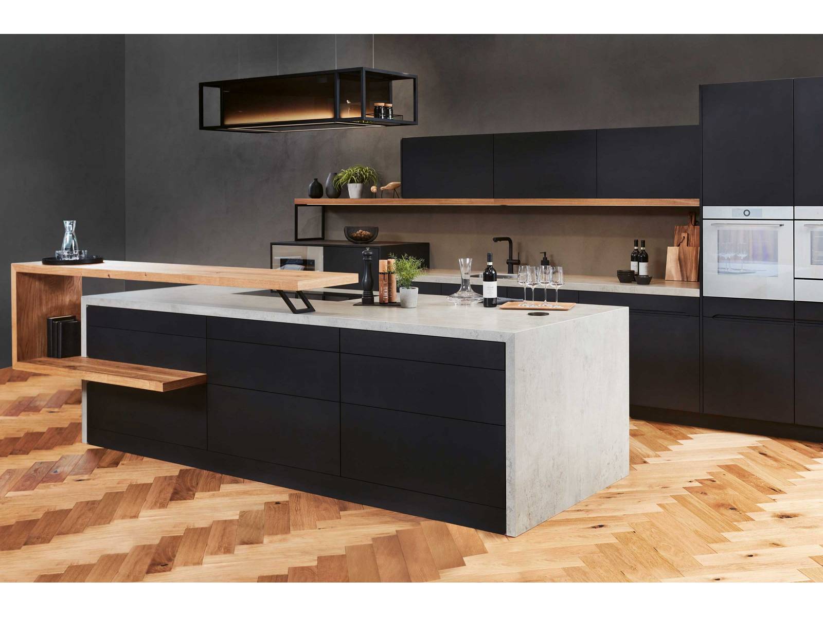 Die Küche der hauseigenen Linie «Infinity» spielt mit offenen und geschlossenen Elementen. Die schwarzen Hängeschränke werden von einem Regalboden aus Holz getragen, der in Form einer Spange vor der Kücheninsel seine Wiederholung findet. Eine betongraue Arbeitsplatte vollendet das neue Modell, das ganz auf Wunsch und nach Mass gefertigt wird. Foto: Studiojeker GmbH. SABAG.