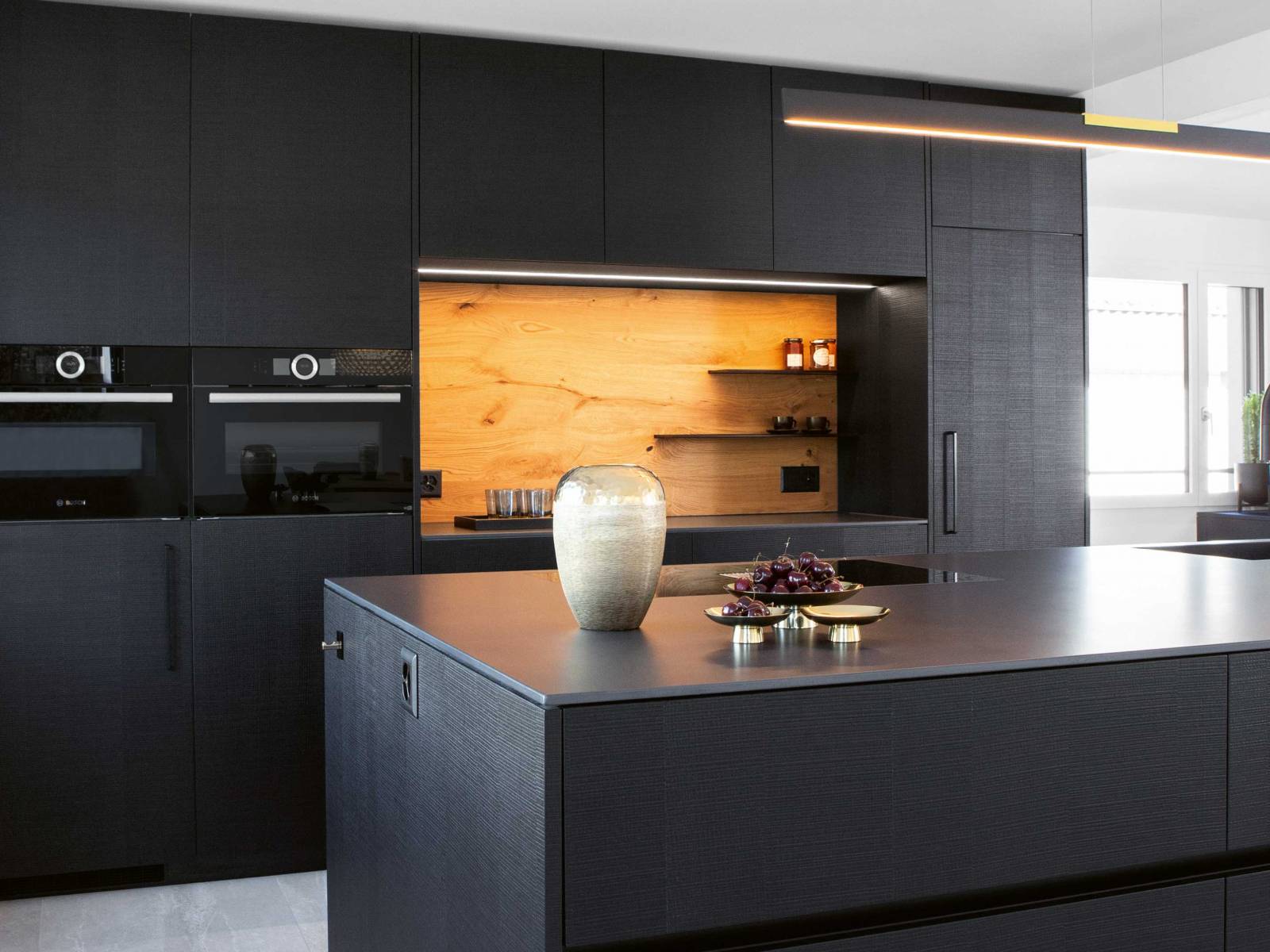 Das sägerohe, schwarz gebeizte und lackierte Eichenfurnier der Küchenfronten gibt jedem Zuhause eine ganz eigene Note und sorgt für ein besonderes haptisches Erlebnis. Während Küchenarmatur und Spülbecken ebenfalls in Schwarz gehalten sind, lockert die Nischenrückwand in heller Eiche das Ensemble auf. Foto: Klaus Andorfer. ELBAU.
