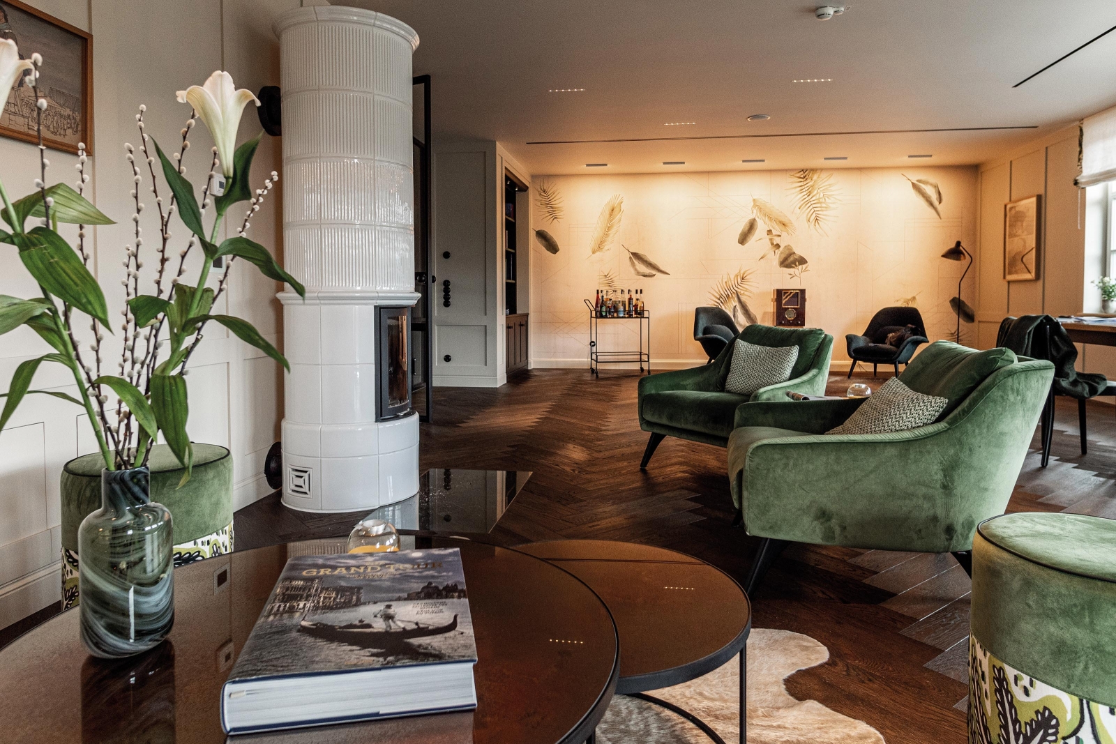 Im Erdgeschoss liegt der grosse Wohn-/Essbereich. Hier befindet sich auch eine kleine Bibliothek mit Barwagen. Farblich abgestimmte Polstermöbel laden zum Lesen und Verweilen ein. Hingucker ist die Tapete «Phytogenesis» mit Blättermotiven von Wall & Deco. Die davor stehenden Loungesessel «Swoon» sind von Fredericia. Die grünen Sessel im Bild vorne sind von IP Design, Modell «Flow Lounge Chair».