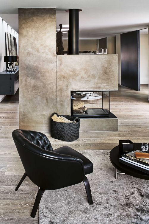 Das Cheminée fungiert als Raumteiler. Je nach Lichteinfall leuchtet es in Gold- und Bronzetönen. Der kapitionierte Ledersessel «Winston», der Salontisch «Torii», beide Minotti, und der Kaminkorb sind ein stimmiges Ensemble in Schwarz.